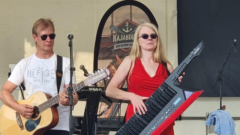 Two musicians performing on stage: a man in a white T-shirt and sunglasses playing an acoustic guitar, and a woman in a red dress playing a keytar behind him.