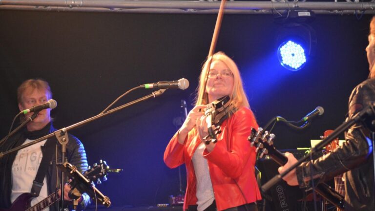 Live music performance with three musicians on stage: a woman in a red jacket playing a violin center stage, flanked by guitarists singing into microphones under blue stage lighting.