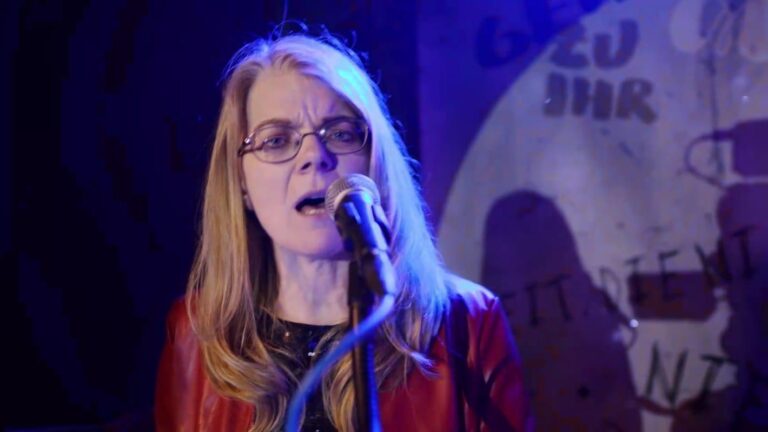Woman with glasses singing into a microphone on a dimly lit stage, wearing a red jacket under blue lighting.