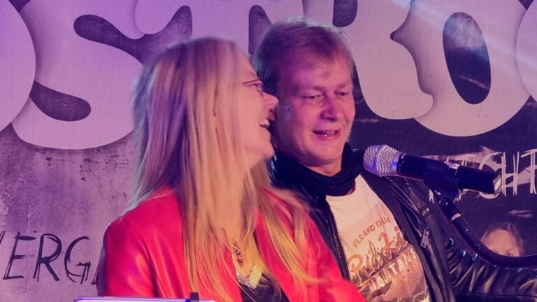 Man and woman singing into a microphone on a stage with a colorful backdrop, smiling at each other.