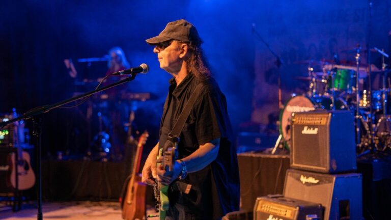 Male guitarist wearing a cap and sunglasses performing on stage with a microphone in front of him, blue stage lighting in the background.