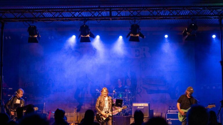 Band performing live on stage under blue lighting; guitarist center, drummer in back, bassist right, audience silhouettes in foreground.