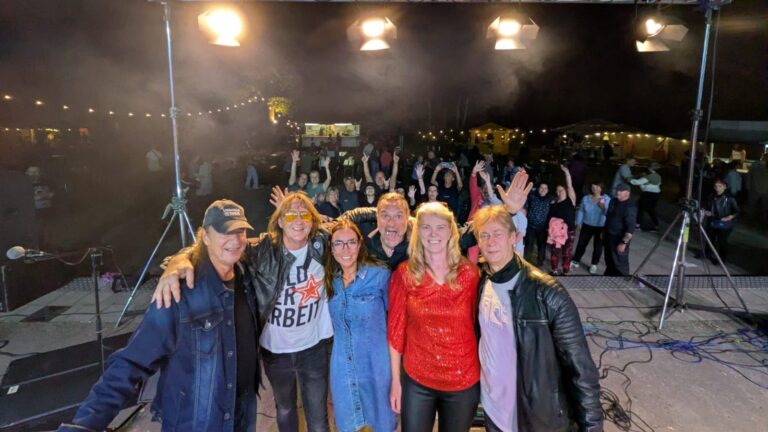 Group of six performers posing for a selfie on a stage at night, crowd cheering with hands raised behind them under string lights.