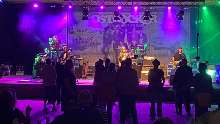 Rock band on stage performing to a standing audience, with purple and green stage lights and a banner in the backdrop.